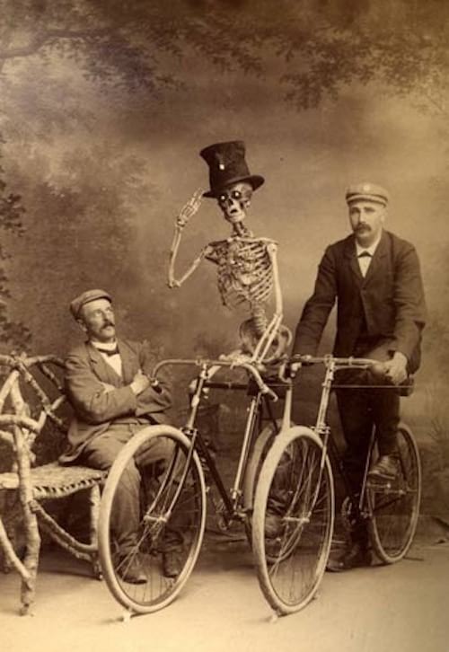 Esqueleto en bicicleta junto a dos hombres con uno sentado y el otro en otra bicicleta