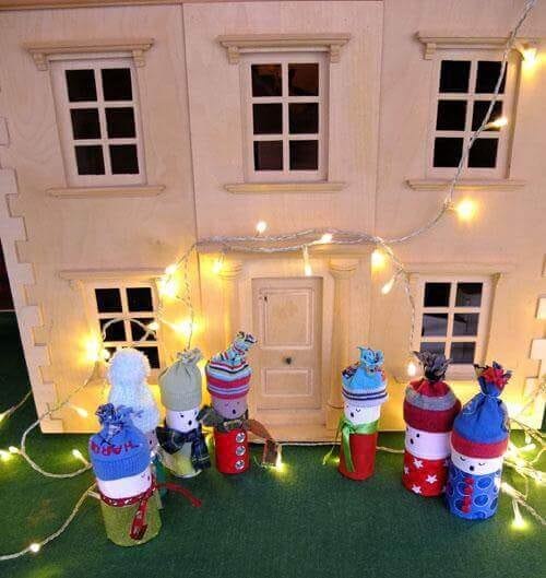 6 muñecos de nieve hechos con rollos de papel higiénico cantando villancicos frente a una casa