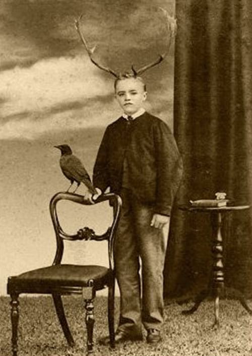 Nen jove amb banyes de cérvol al cap mirant a un costat sostenint una cadira al seu costat i un corb a sobre