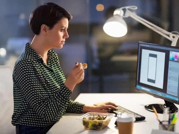 Una mujer come su comida frente a la computadora.